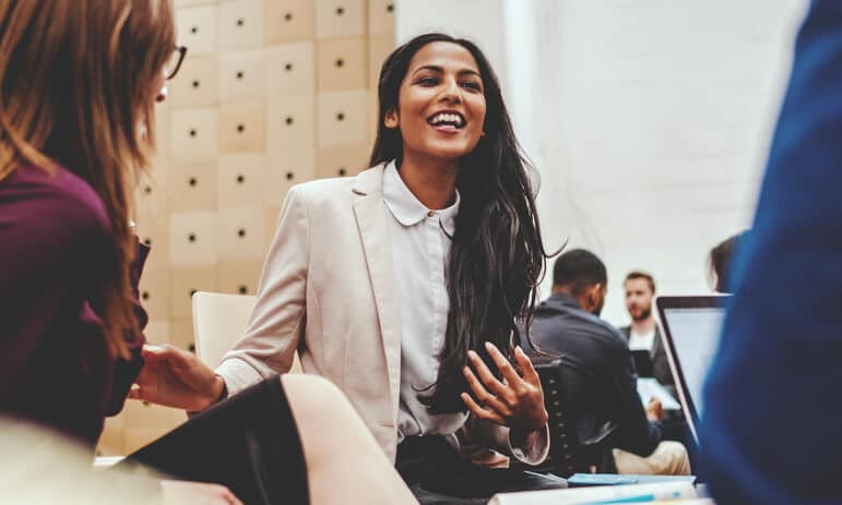 A woman with long, dark hair, wearing a light-colored blazer over a white shirt, is smiling and talking with two other people in a modern office setting. As a certified professional accountant, she appears to be engaged and enthusiastic.