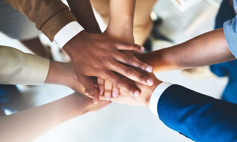 A group of people, including certified professional accountants, place their hands together in a central stack, symbolizing teamwork and unity. The hands are of varying skin tones and belong to individuals dressed in both casual and business attire. The background is blurred, emphasizing the hands.