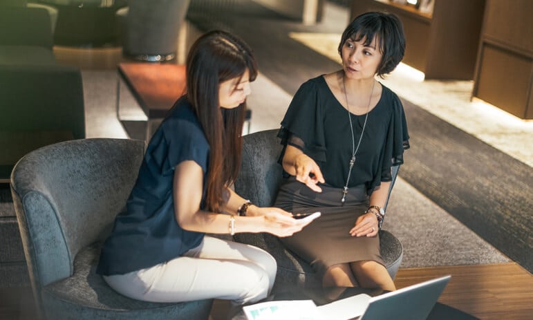 Two women are sitting on cushioned chairs in a modern, well-lit office space. One woman is holding a smartphone and gesturing towards it while the other woman, a certified professional accountant with a pen in hand, is attentively listening and looking at the phone. A laptop is open on the table in front of them.