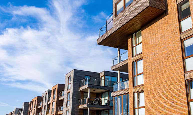 A row of modern apartment buildings with wooden and brick facades under a blue sky with scattered clouds. Each building features multiple balconies with glass railings, and large windows that reflect the sky. The architecture is contemporary and clean—ideal for tax accountants seeking a serene urban retreat.