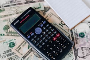 A black scientific calculator and a small, blank notepad rest on top of scattered U.S. dollar bills, hinting at the meticulous work of CPAs. The calculator displays various buttons for mathematical functions, and the money includes denominations such as $10, $20, and others.