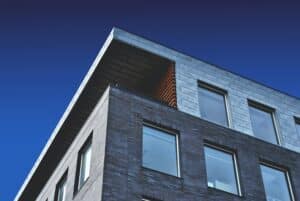 A modern, multi-story building with gray brick and metal siding is shown from a low angle. The building, possibly housing tax accountants, has several large, rectangular windows and features a sharp, angular roof against a clear blue sky.