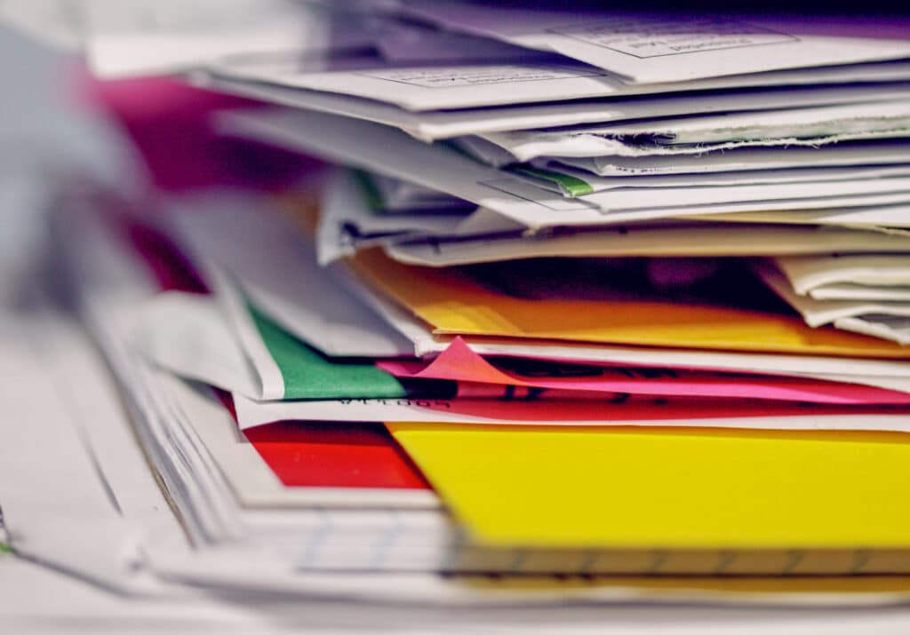 A close-up image of a stack of various papers, folders, and envelopes in disarray, likely the workspace of certified professional accountants. The documents are of different colors, including yellow, red, and green, creating a vibrant and cluttered appearance. The focus is shallow, blurring some edges of the papers.